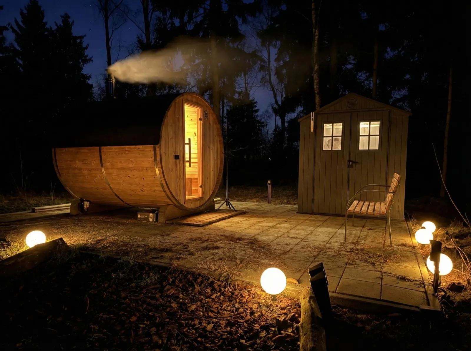 Houten barrelsauna met dampende schoorsteen in de avond, naast een tuinhuis